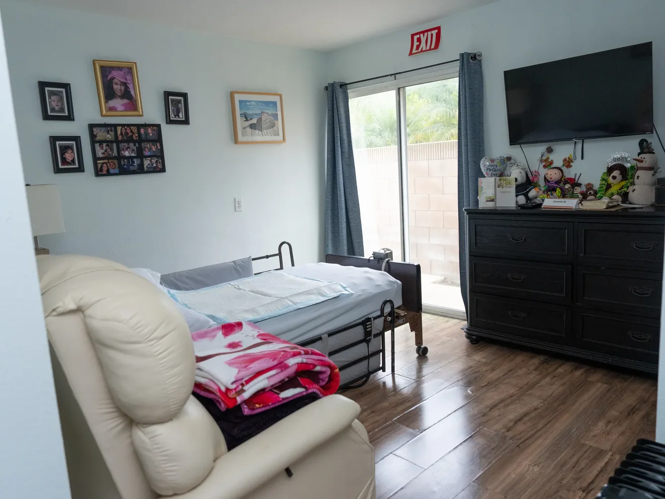 Resident bedroom with hospital bed, wood dresser, and sliding door outdoors.